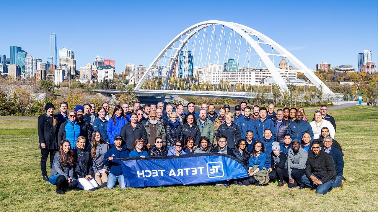 A large group of employees holding a Tetra Tech banner at a park with a city skyline and bridge in the background